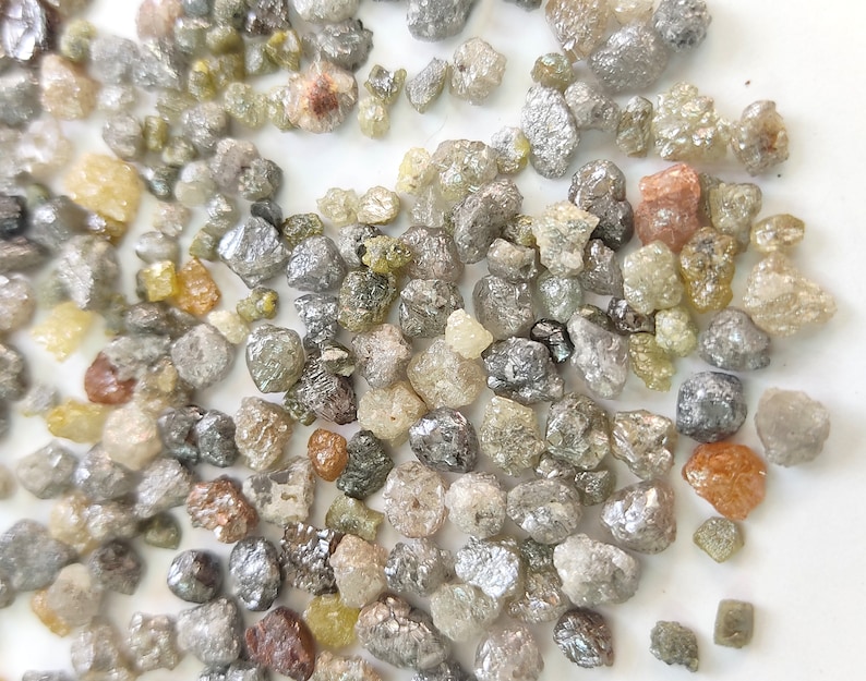 Puede incluir: Una colecci&oacute;n de peque&ntilde;os diamantes en bruto, grises y verdes. Los diamantes est&aacute;n dispersos sobre una superficie blanca.