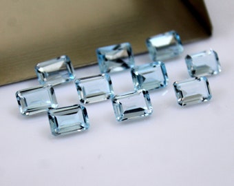 May include: A collection of twelve light blue, rectangular, faceted gemstones. The gemstones are arranged in a staggered pattern on a white surface.