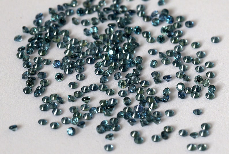 Op de afbeelding: Een strooiing van teal blauwe edelstenen op een wit oppervlak. De edelstenen zijn rond en hebben een facetgeslepen.