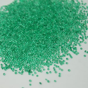 May include: A large pile of small, round, green gemstones. The gemstones are scattered on a white surface.