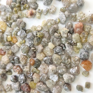 Puede incluir: Una colecci&oacute;n de peque&ntilde;os diamantes en bruto, grises y amarillos. Los diamantes est&aacute;n dispersos sobre una superficie blanca.