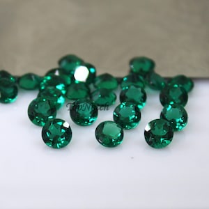 May include: A collection of round, faceted, green gemstones. The gemstones are scattered on a white surface.