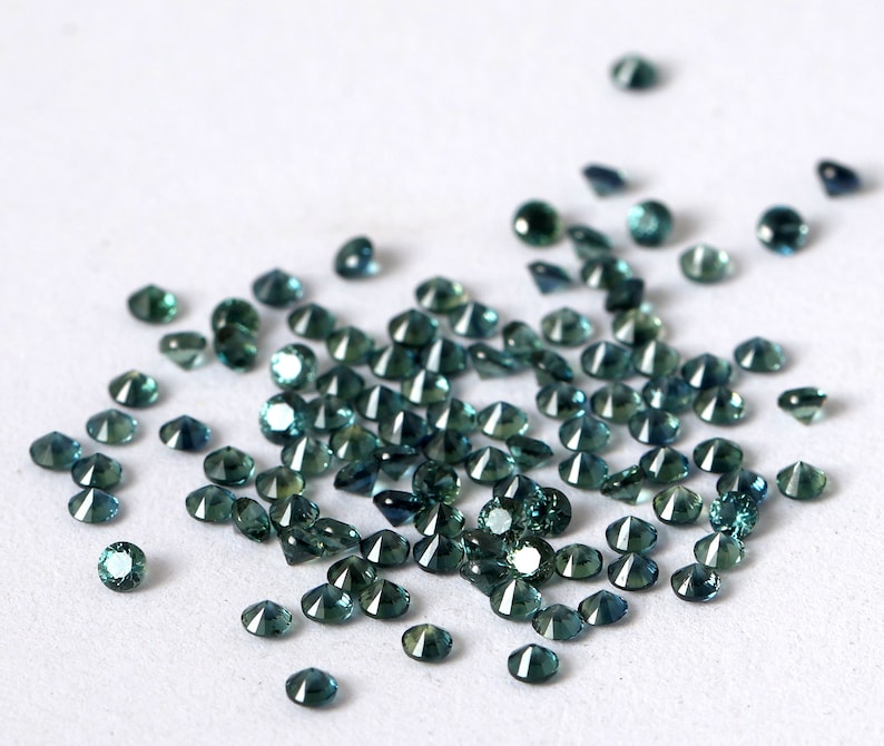 Op de afbeelding: Een verzameling losse teal blauwe edelstenen, waarschijnlijk saffieren, verspreid over een wit oppervlak. De edelstenen zijn gefacetteerd en hebben een briljante glans.