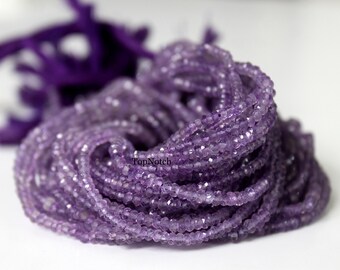 May include: A strand of purple amethyst beads, faceted and round, with a close-up view of the beads. The beads are arranged in a pile, showing the texture and color of the amethyst.