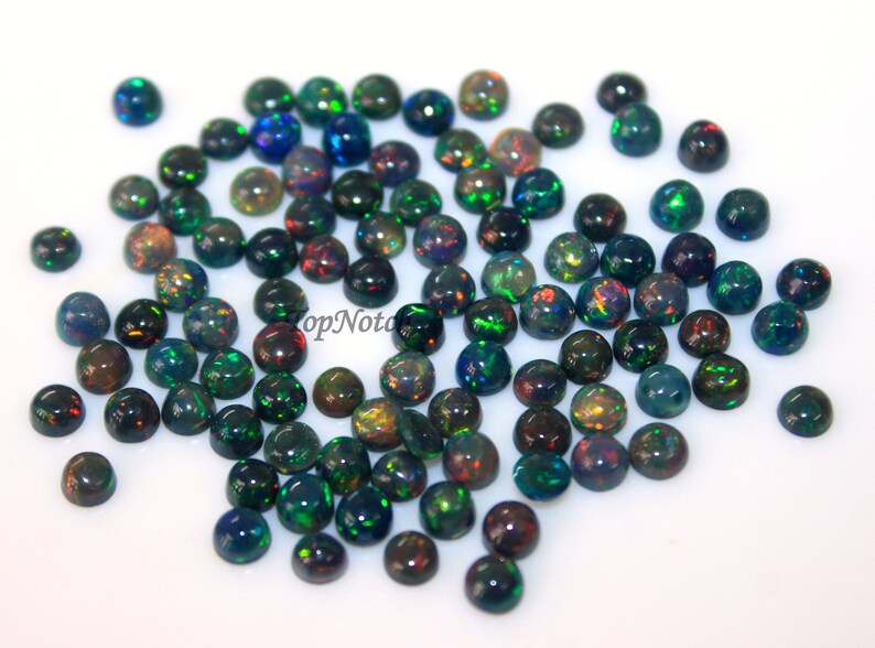 May include: A collection of round, black opal cabochons with iridescent green, blue, and red flashes. The opals are scattered on a white surface.