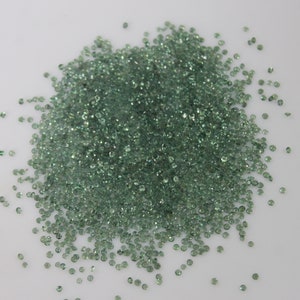 Op de afbeelding: Een grote hoop kleine, ronde, groene edelstenen. De edelstenen zijn verspreid over een wit oppervlak.
