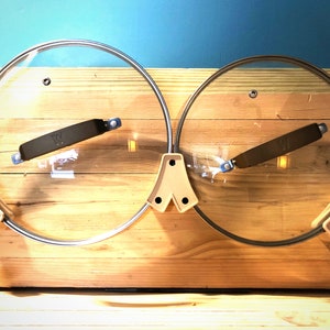 Puede incluir: Dos tapas de ollas de vidrio transparente con bordes de acero inoxidable y asas negras se exhiben en un soporte de madera. El soporte tiene un marco beige claro y está sobre un fondo azul. Las tapas son redondas y tienen un pequeño pomo de metal.