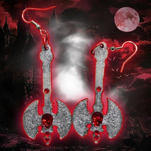 May include: A pair of silver-colored axe-shaped earrings with red accents. Each earring features a skull at the blade's center and a red gem. The earrings are set against a dark, gothic-themed background with a full moon.