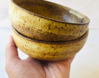 May include: Two handmade ceramic bowls. The bowls are stacked, with a speckled, earthy yellow-brown glaze. The rims are a darker brown. The bowls are held in a hand, with a light background.
