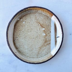 May include: A handmade ceramic plate with a speckled beige interior and a dark brown, textured rim. The plate has a unique, organic shape and a glossy white section on the side. This rustic plate is perfect for serving food.