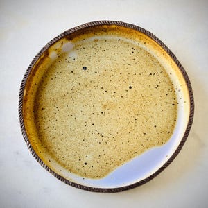 May include: A handmade ceramic plate with a speckled tan interior and a dark brown, textured rim. The plate has a unique, irregular shape and a light blue accent on one side. The plate is suitable for serving food.