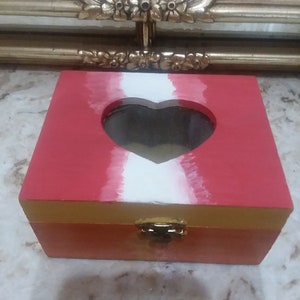 May include: A red wooden box with a heart-shaped cutout and a gold trim. The box has a white heart design on the front and a gold latch.
