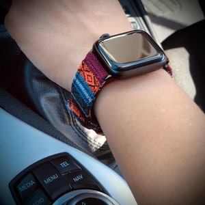 May include: A black smartwatch with a colorful patterned band featuring orange, blue, and purple geometric designs. The watch is worn on a wrist, showcasing the band's intricate details and the watch's sleek, modern design.