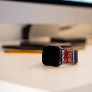 May include: A black smartwatch with a colorful woven band featuring a geometric pattern in red, orange, white, and blue. The watch is displayed on a white surface, with a blurred background.