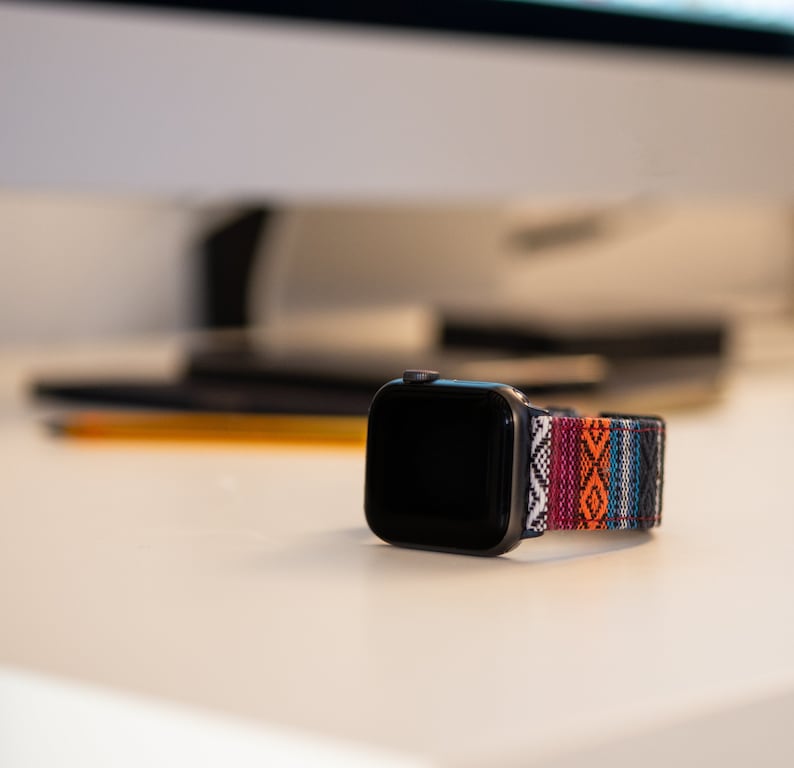 May include: A black smartwatch with a colorful woven band featuring a geometric pattern in red, orange, white, and blue. The watch is displayed on a white surface, with a blurred background.