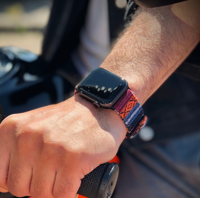 May include: Black smartwatch with a colorful woven band featuring a geometric pattern in orange, red, blue, and white. The watch is worn on a wrist, with the band secured. The watch is a modern accessory.