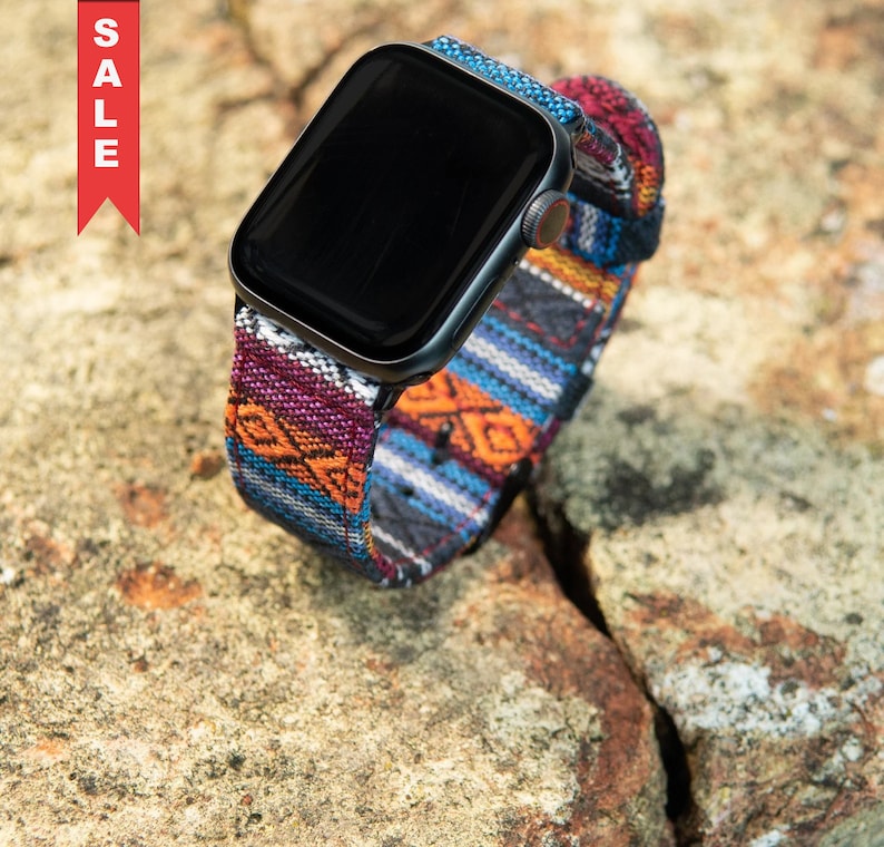 May include: A black smartwatch with a colorful woven band featuring a geometric pattern in shades of blue, orange, purple, and white. The word "SALE" is printed vertically on a red tag in the upper left corner.
