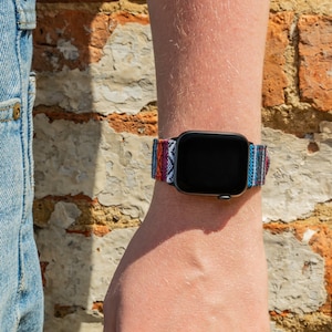 May include: A black smartwatch with a colorful woven band featuring a geometric pattern in shades of red, blue, white, and orange. The watch is worn on a wrist, with a brick wall in the background.