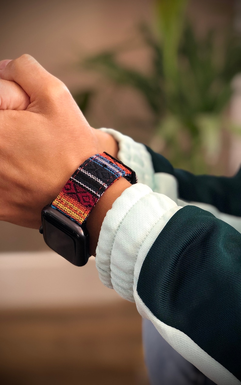 May include: Black smartwatch with a colorful woven band. The band features a geometric pattern in red, orange, blue, and white. The watch is worn on a wrist, with a green and white jacket in the background.