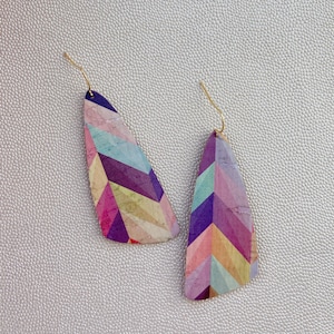 May include: A pair of geometric patterned earrings with a light purple, pink, blue, and yellow colour scheme. The earrings are made of cork and have a gold wire.