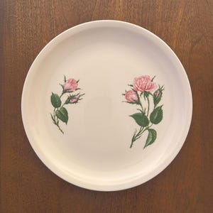 Op de afbeelding: Een rond, gebroken wit keramieken bord met twee afzonderlijke bloemillustraties. Links een enkele roze rozenknop met groene bladeren. Rechts een groep van twee roze rozen en een knop met groene bladeren en doornige stengels.