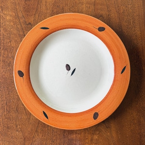 May include: A round ceramic plate featuring a wide, textured orange rim and a smooth, off-white center. The design includes small, dark brown, leaf-like motifs scattered across both the orange border and the central white area of the plate.