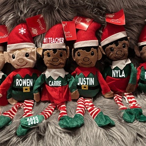May include: Six plush elf dolls with red and green outfits, each with a name on the front. The dolls have red hats and striped legs. One doll says "#1 TEACHER" and another has the year "2023" on its foot.