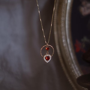 Può includere: Una collana a catena d'oro con un ciondolo a forma di cuore rosso. Il ciondolo è circondato da un cerchio di diamanti più piccoli. La collana è appesa su uno sfondo scuro.