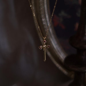May include: A gold necklace with a small cross pendant featuring red gemstones. The necklace is hanging from a vintage-style ornate mirror.