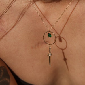 May include: A gold necklace with a green gemstone pendant and a sword charm. The sword charm is adorned with small clear gemstones.