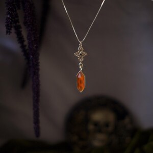 Op de afbeelding: Een zilveren ketting met een decoratieve hanger met een kleine, oranje kristal. De hanger heeft een gedetailleerd, sierlijk ontwerp met een kruisvorm en kleine zwarte accenten. De ketting hangt tegen een donkere achtergrond.