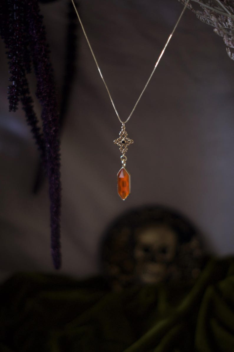 Op de afbeelding: Een zilveren ketting met een decoratieve hanger met een kleine, oranje kristal. De hanger heeft een gedetailleerd, sierlijk ontwerp met een kruisvorm en kleine zwarte accenten. De ketting hangt tegen een donkere achtergrond.