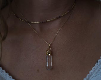 May include: A gold pendant necklace featuring a clear crystal quartz point with gold accents. A delicate beaded necklace in shades of pink and gold sits above it. The necklaces are worn on a person wearing a white top.