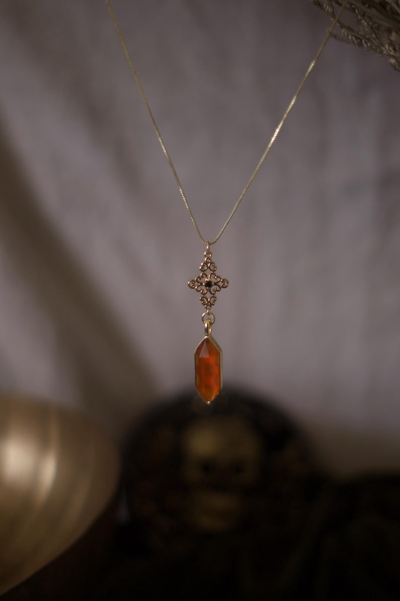 Op de afbeelding: Een goudkleurige ketting met een hanger met een kruisvormig ontwerp met zwarte accenten en een lange, oranje kristal. De ketting hangt tegen een zachte, vage achtergrond, waardoor de ingewikkelde details en warme kleuren van de hanger worden benadrukt.