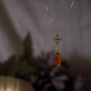 Op de afbeelding: Een goudkleurige ketting met een hanger met een decoratief filigraanontwerp en een gefacetteerde, oranje-rode edelsteen. De ketting hangt tegen een vage achtergrond, waardoor de details van de sieraden worden benadrukt.
