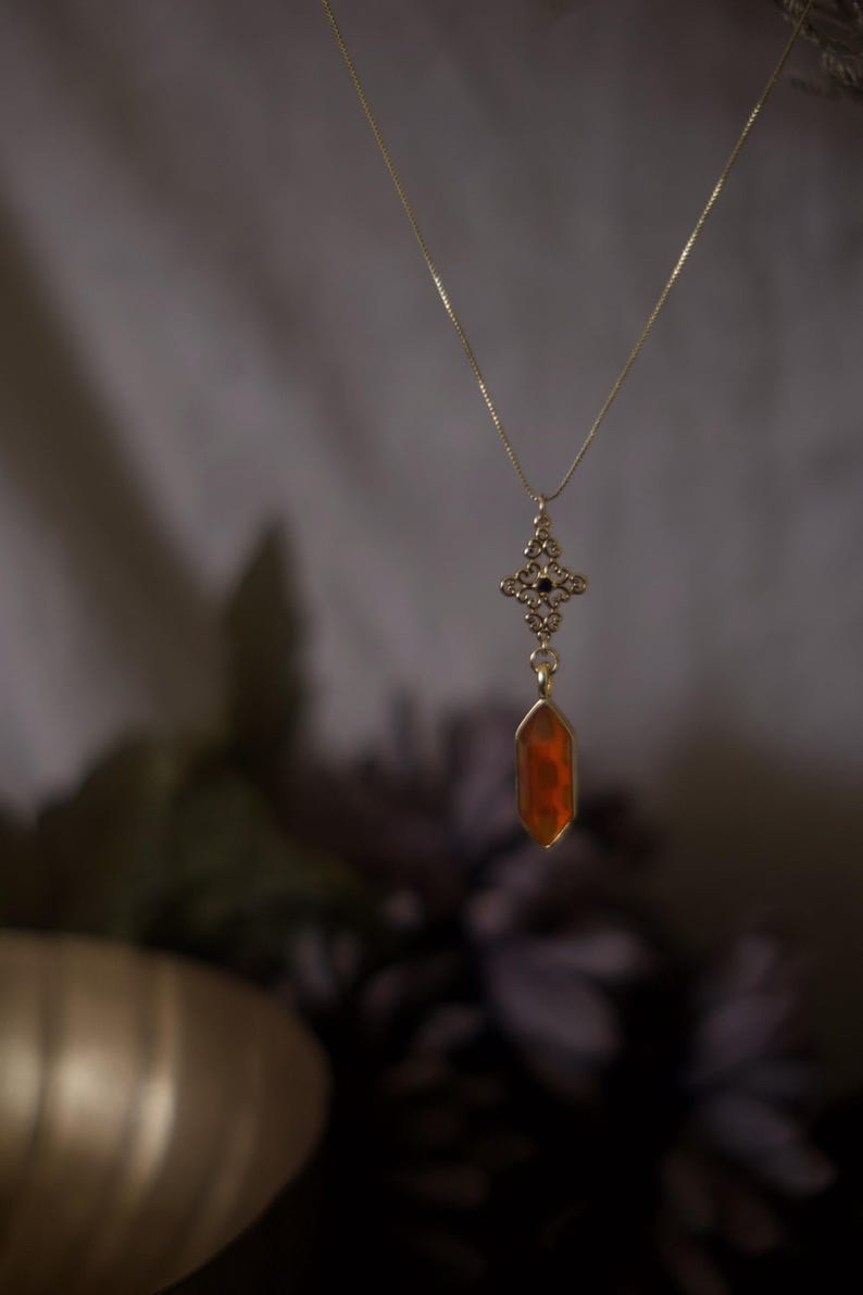 Op de afbeelding: Een goudkleurige ketting met een hanger met een decoratief filigraanontwerp en een gefacetteerde, oranje-rode edelsteen. De ketting hangt tegen een vage achtergrond, waardoor de details van de sieraden worden benadrukt.