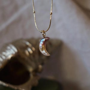 Pode incluir: Um delicado colar dourado com um pingente de lua crescente. O pingente tem um acabamento perolado com tons iridescentes de rosa, roxo e dourado. A corrente é uma corrente fina dourada com pequenas contas decorativas.