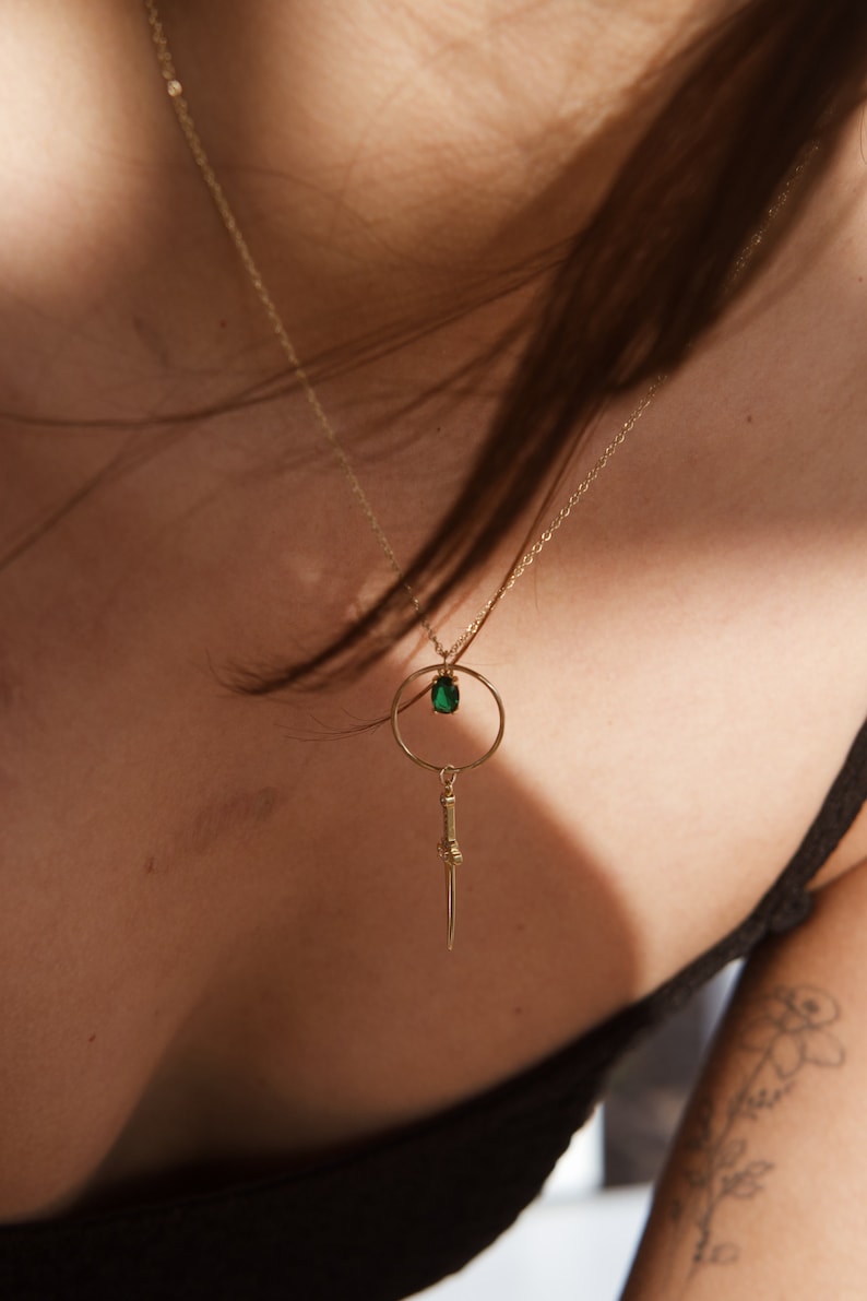 May include: A gold necklace with a green gemstone pendant and a dagger charm hanging from a circle.