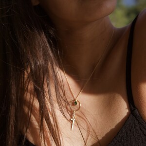 May include: A woman wearing a black tank top with a gold chain necklace featuring a small green stone and a cross pendant.