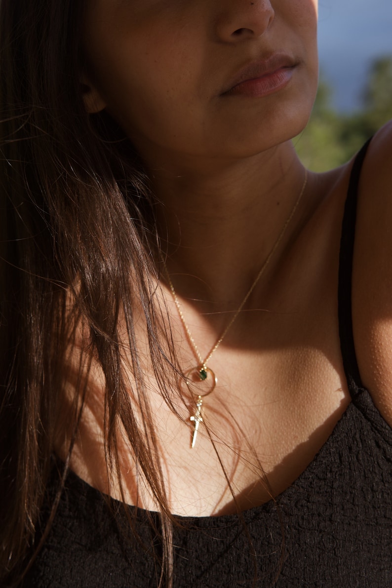 May include: A woman wearing a black tank top with a gold chain necklace featuring a small green stone and a cross pendant.