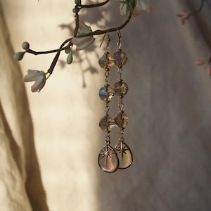 May include: A pair of long dangle earrings featuring three stacked, faceted, iridescent glass beads in a warm amber tone, finished with a teardrop-shaped charm at the base that displays a delicate, etched starburst design in the center.