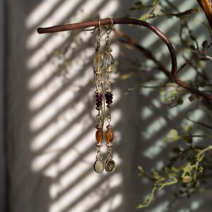 Puede incluir: Un par de pendientes largos y colgantes con un dise&ntilde;o de metal dorado. Los pendientes presentan una variedad de cuentas en tonos amarillos, naranjas y rojos. Cuelgan de una rama marr&oacute;n, con un fondo borroso.