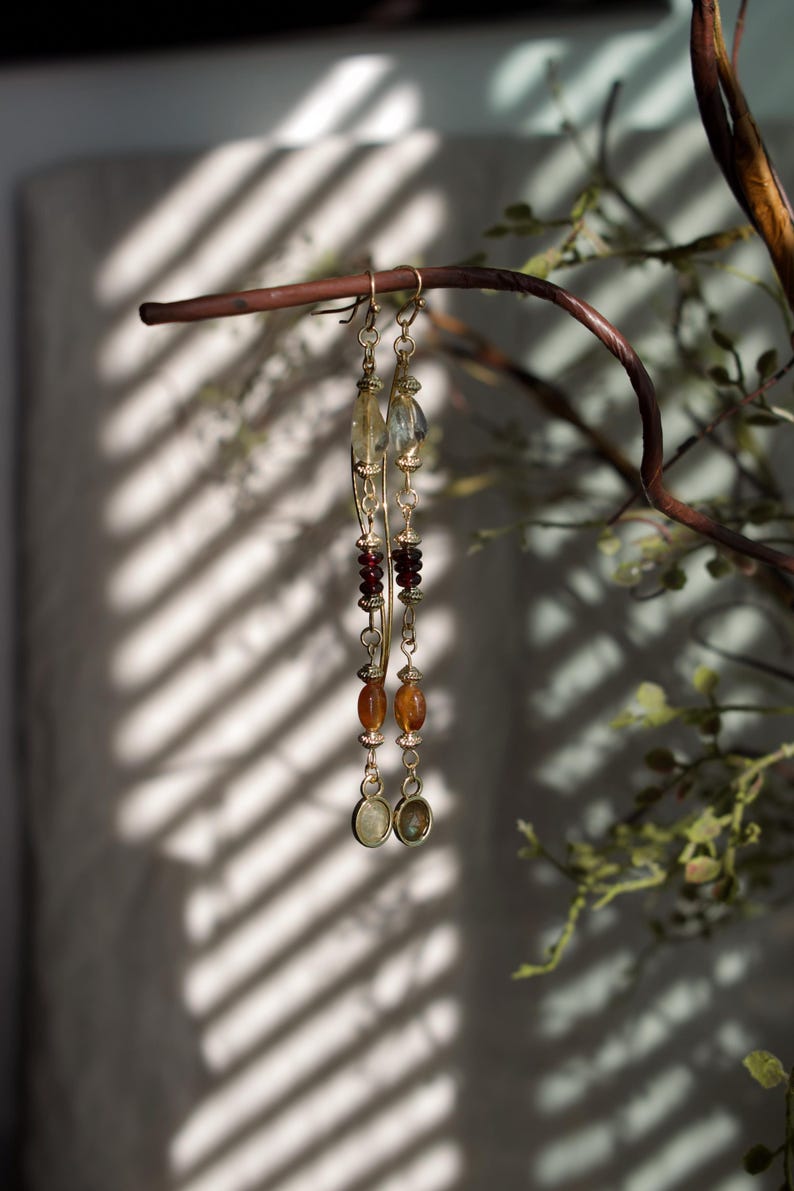 Puede incluir: Un par de pendientes largos y colgantes con un dise&ntilde;o de metal dorado. Los pendientes presentan una variedad de cuentas en tonos amarillos, naranjas y rojos. Cuelgan de una rama marr&oacute;n, con un fondo borroso.