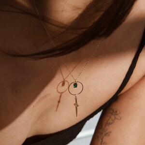 May include: A gold necklace with a green gemstone pendant and a dagger charm. The necklace is worn by a person with a tattoo on their arm.
