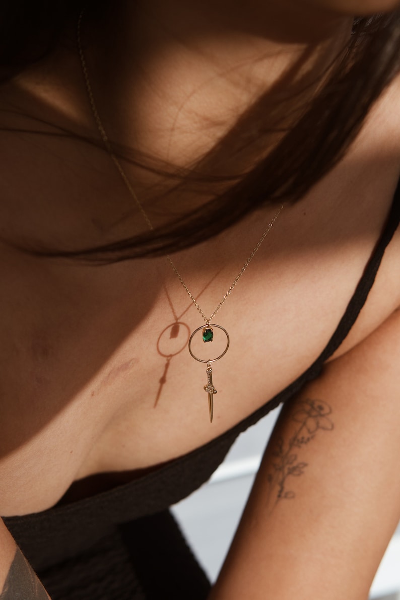 May include: A gold necklace with a green gemstone pendant and a dagger charm. The necklace is worn by a person with a tattoo on their arm.