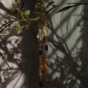 Puede incluir: Pendientes colgantes dorados con una variedad de piedras preciosas, incluyendo cuentas claras, &aacute;mbar y rojo oscuro. Los pendientes cuelgan de una rama con hojas verdes, proyectando sombras en el fondo.