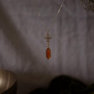 Op de afbeelding: Een goudkleurige ketting met een hanger met een kruisvormig ontwerp en een hangende, oranje, zeshoekige edelsteen. De ketting hangt tegen een zachte, neutrale achtergrond, met gedroogd gebladerte in de rechterbovenhoek.