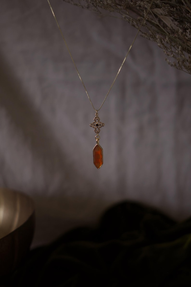 Op de afbeelding: Een goudkleurige ketting met een hanger met een kruisvormig ontwerp en een hangende, oranje, zeshoekige edelsteen. De ketting hangt tegen een zachte, neutrale achtergrond, met gedroogd gebladerte in de rechterbovenhoek.