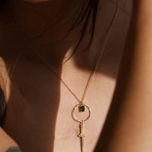 May include: A gold necklace with a green gemstone pendant and a dagger charm. The necklace is on a person's chest.