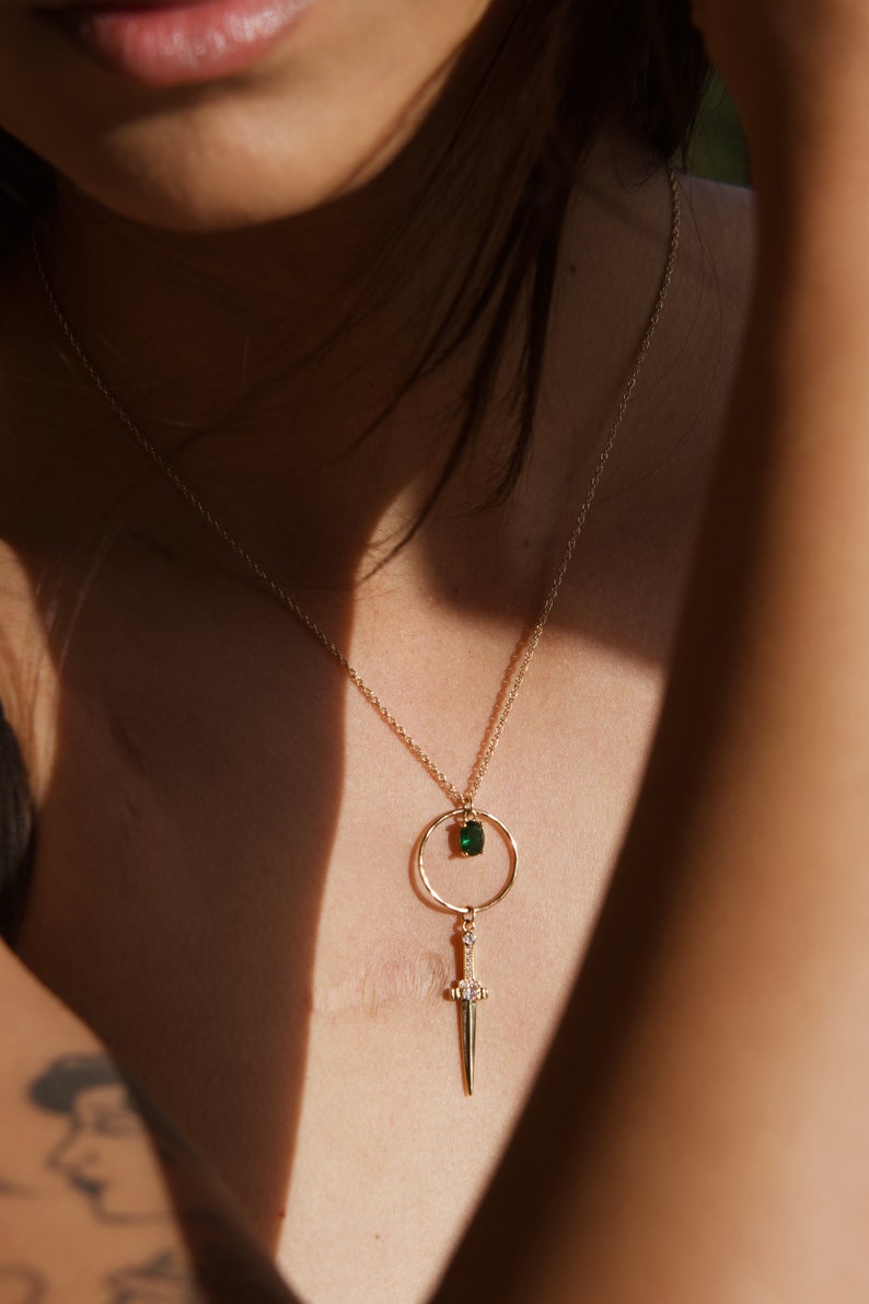 May include: A gold necklace with a green gemstone pendant and a dagger charm. The necklace is on a person's chest.